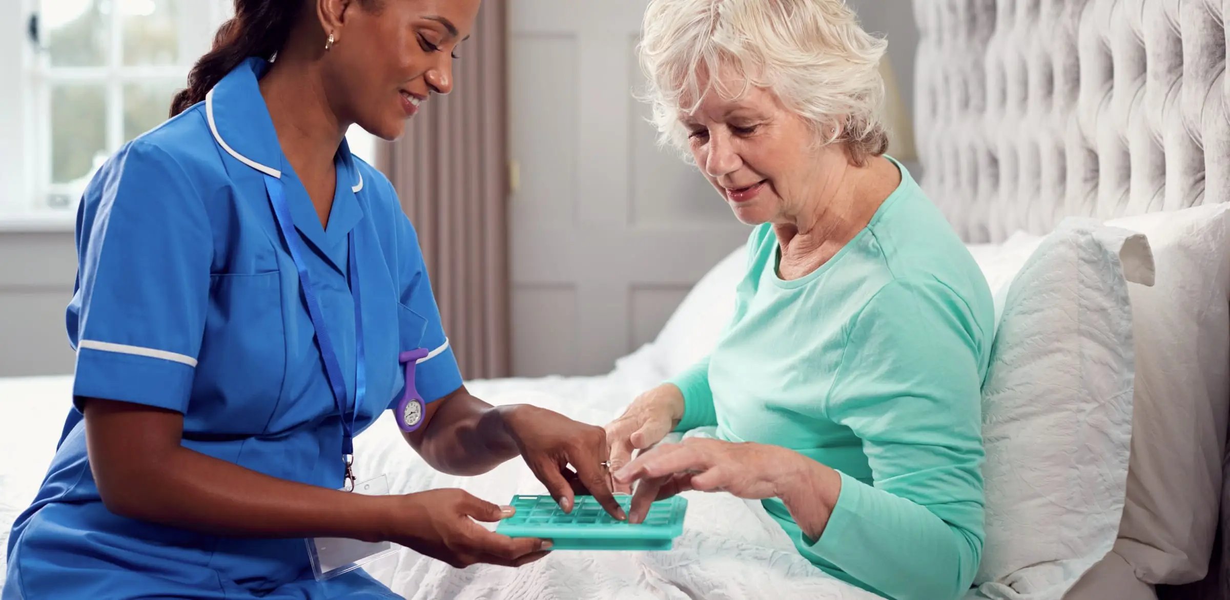 A caregiver sits at the bedside, helping an elderly resident sort her medication into a pill organiser